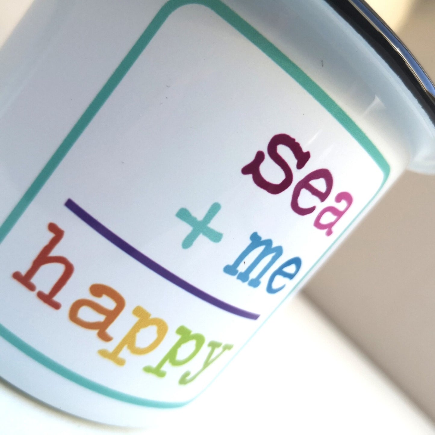 A close up photo of a White enamel mug with a black rim with the following on the front - SEA + ME = HAPPY in rainbow colours