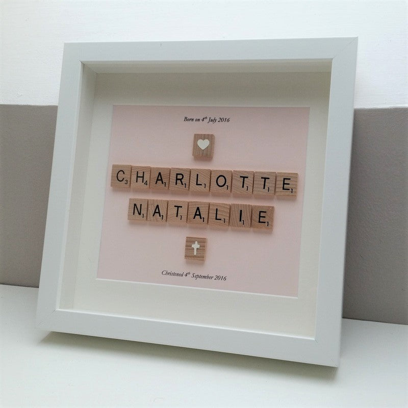 A name in wooden scrabble tiles on a pastel pink background with the date of their birth and their christening above and below. In a white wooden handmade 25x25cm deep box frame.