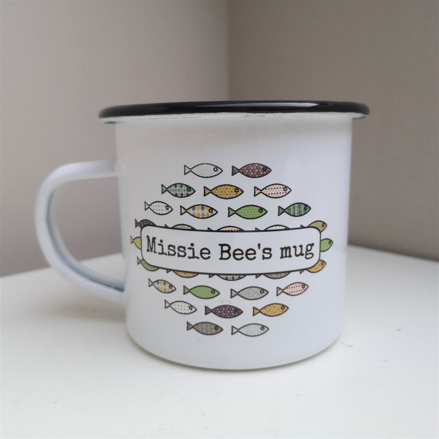 A white enamel mug, with a colourful shoal of fish on the front, overlayed with the owner's name.
