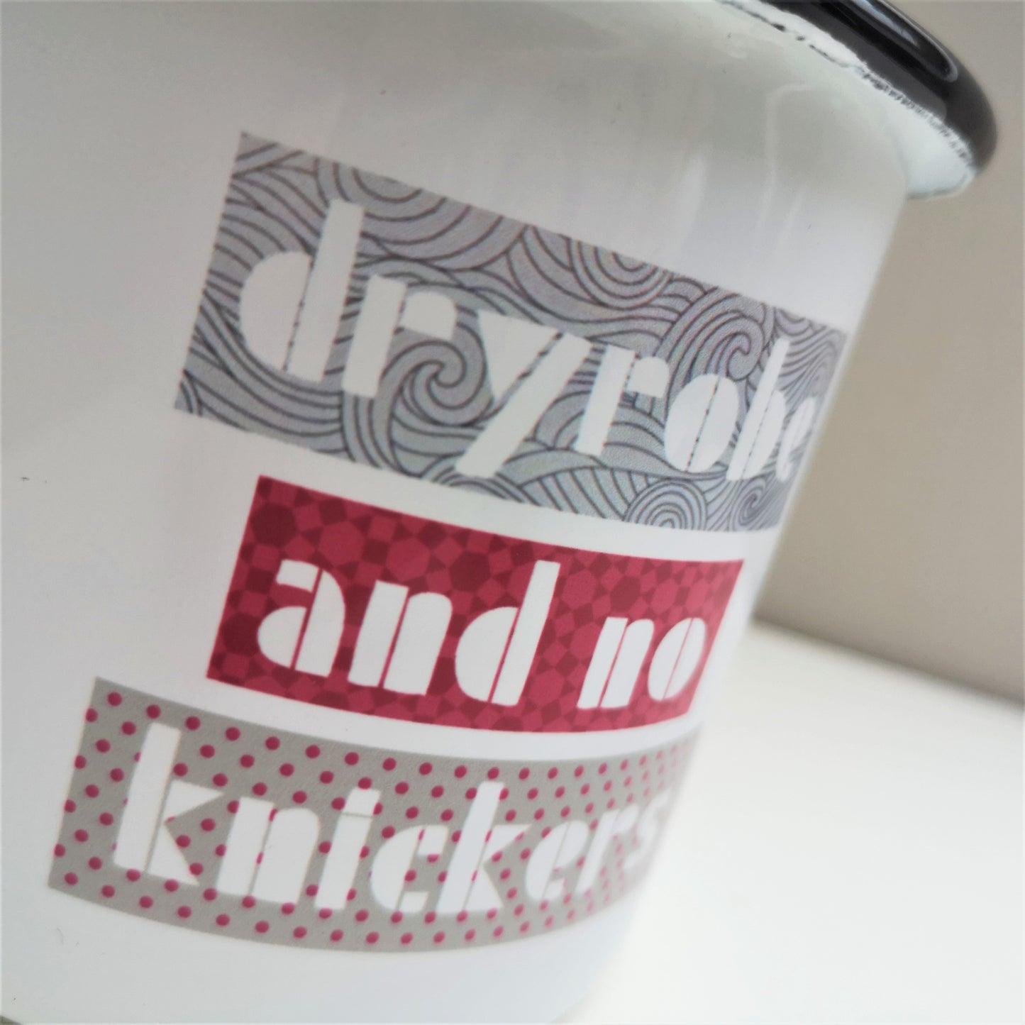 A close up of a white steel enamel mug with DRYROBE AND NO KNICKERS on the front with blocks of greys and dark pinks around the letters