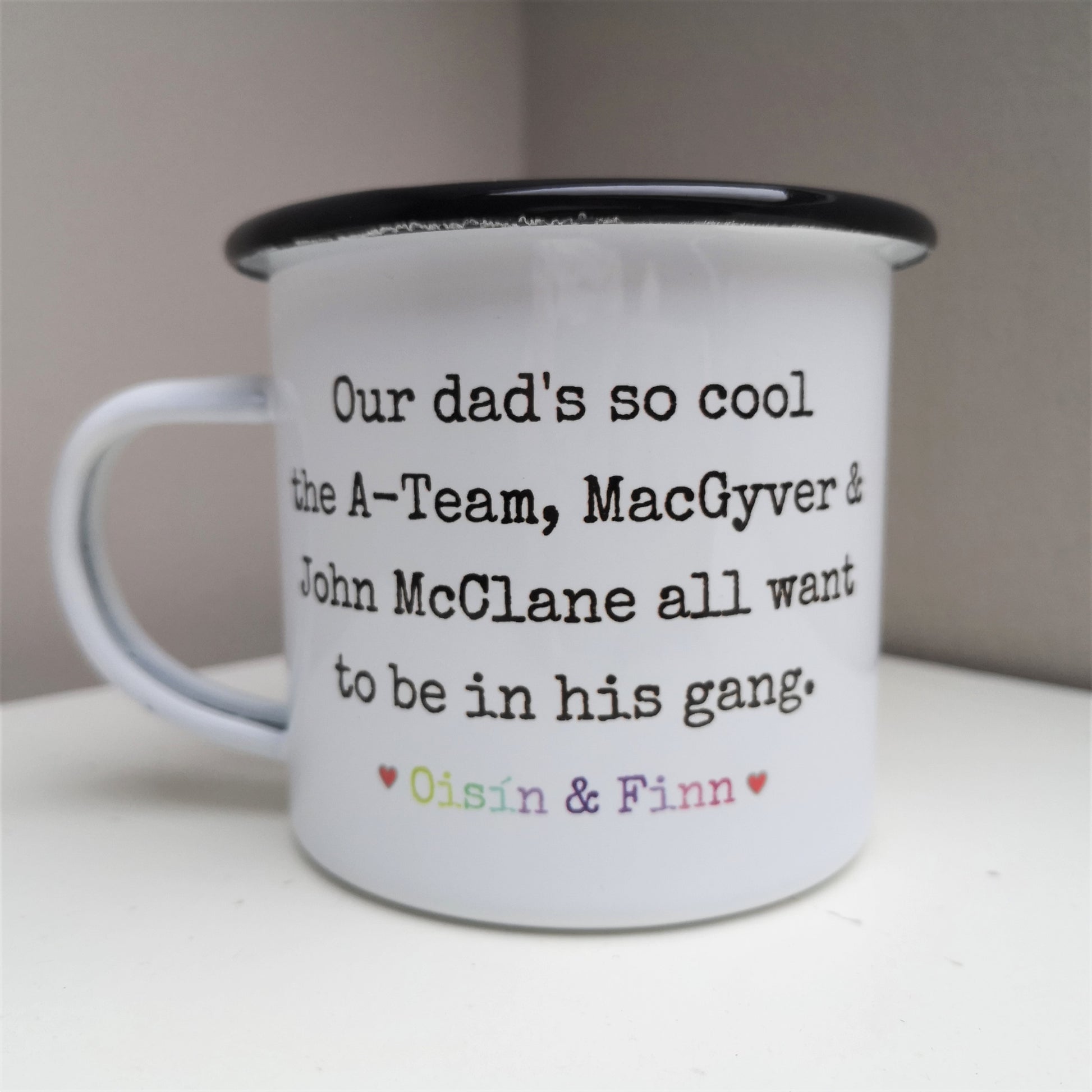 A White enamel mug with a black rim with the following on the front - Our dad's so cool the A-Team, MacGyver and John McClane all want to be in his gang. Below that text is a space for personalised names to go.