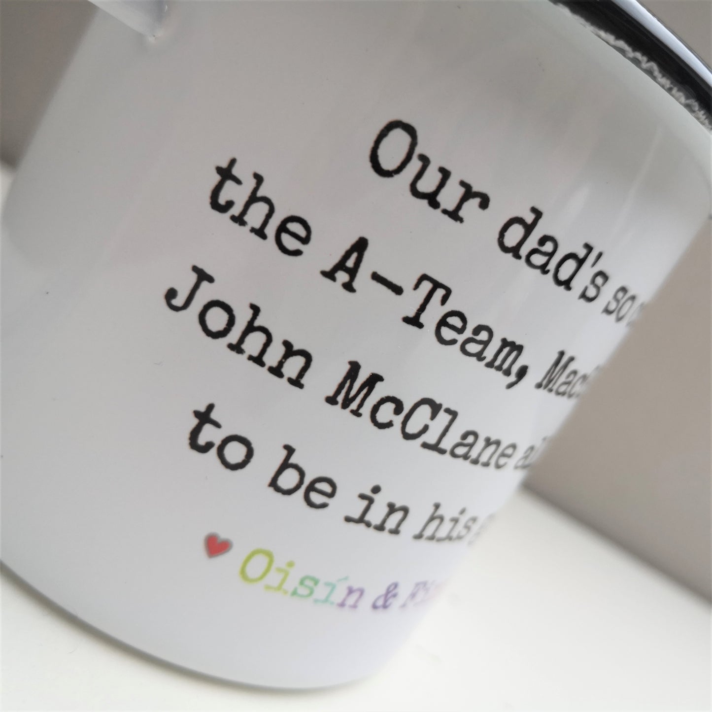 A close up of a White enamel mug with a black rim with the following on the front - Our dad's so cool the A-Team, MacGyver and John McClane all want to be in his gang. Below that text is a space for personalised names to go.