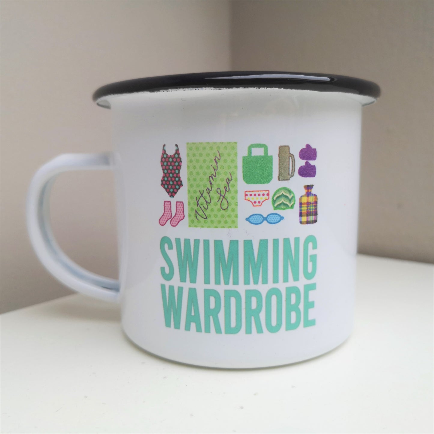 A White enamel mug with a black rim with the following on the front - a mini swimming wardrobe of togs, booties, towel, bag, pants, goggles, swimming cap, flask, gloves and hot water bottle