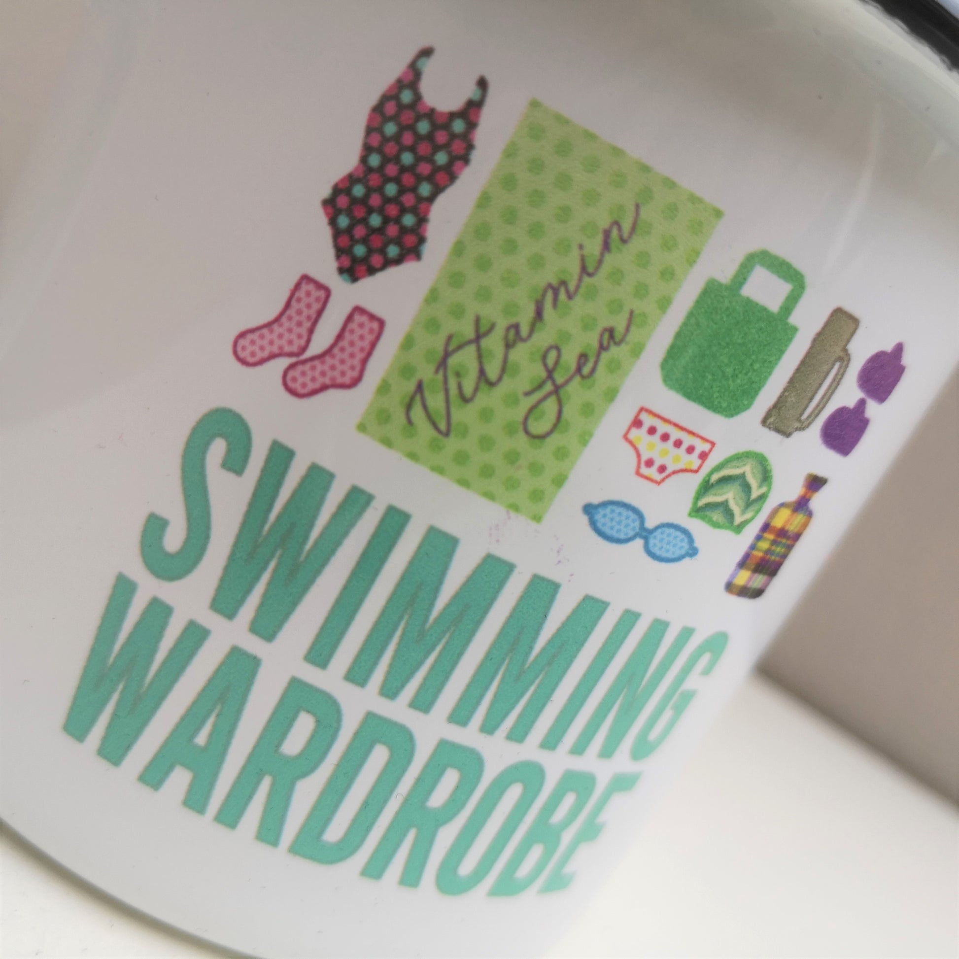 A close up of a White enamel mug with a black rim with the following on the front - a mini swimming wardrobe of togs, booties, towel, bag, pants, goggles, swimming cap, flask, gloves and hot water bottle