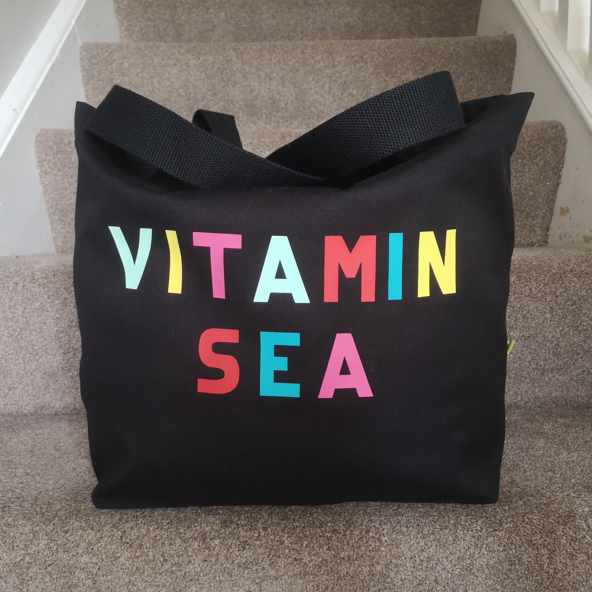 A different view of a black organic cotton tote with toggle closing with VITAMIN SEA in different coloured bright lettering on the front