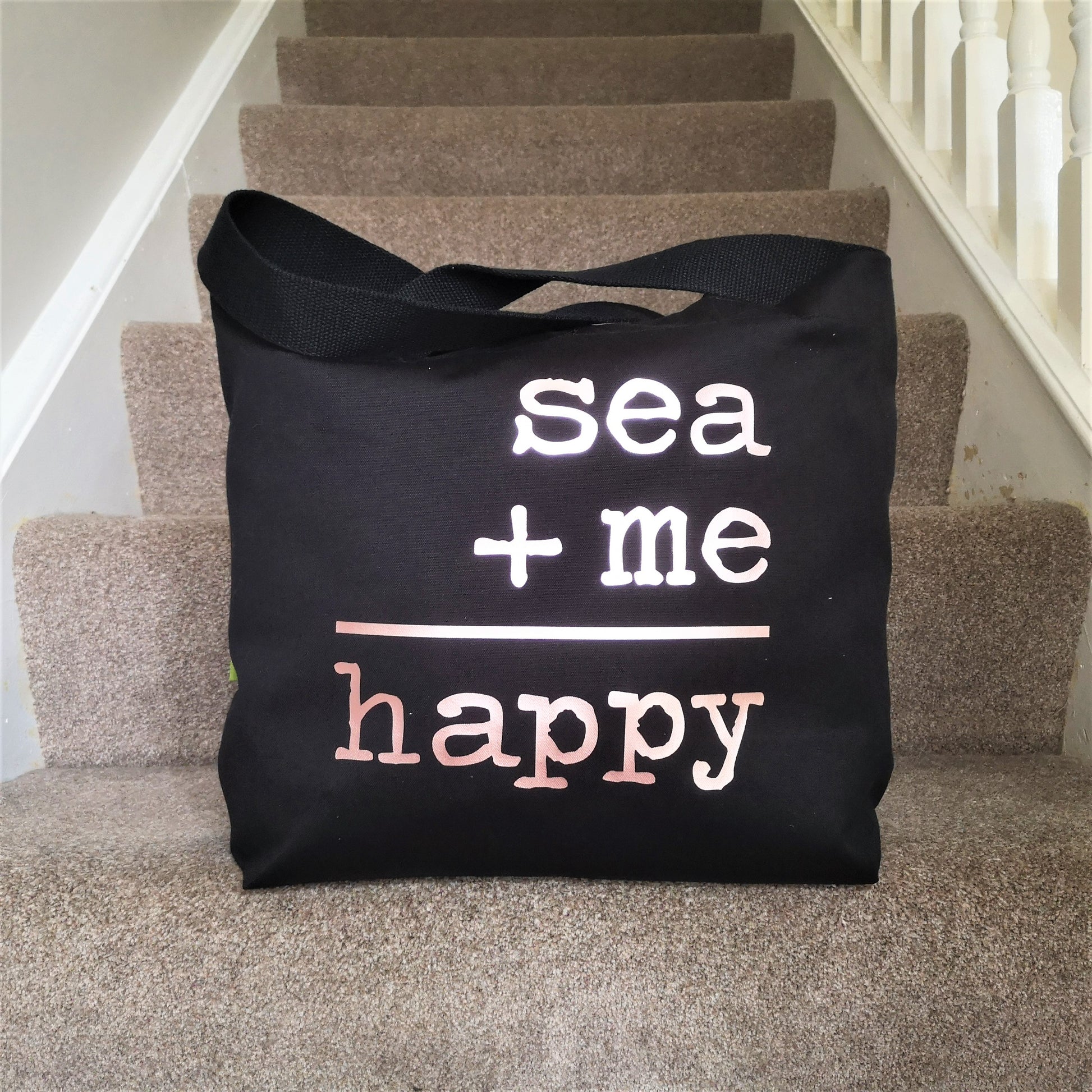 A black organic cotton tote with toggle closing with the following text on the front in white type font - SEA + ME = HAPPY