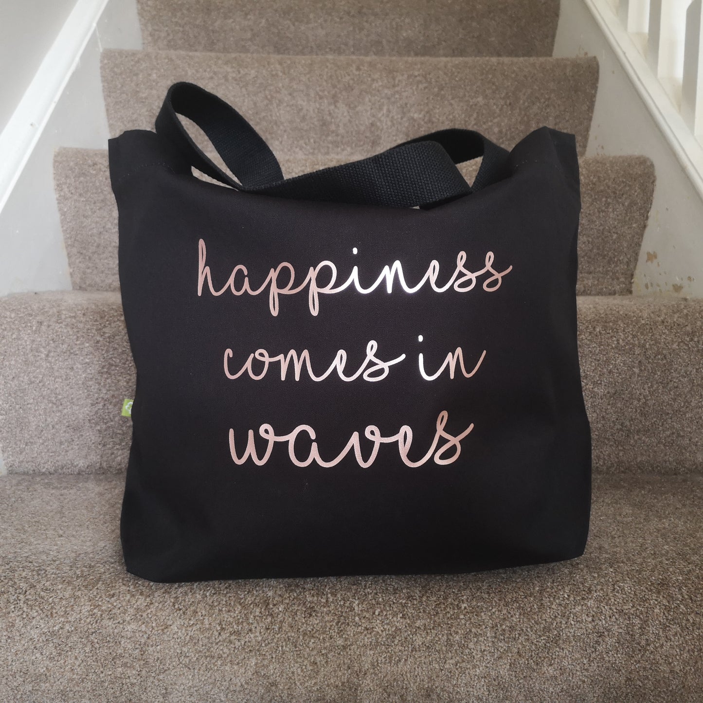 A black organic cotton tote with toggle closing with the following text on the front in rose gold type font - HAPPINESS COMES IN WAVES