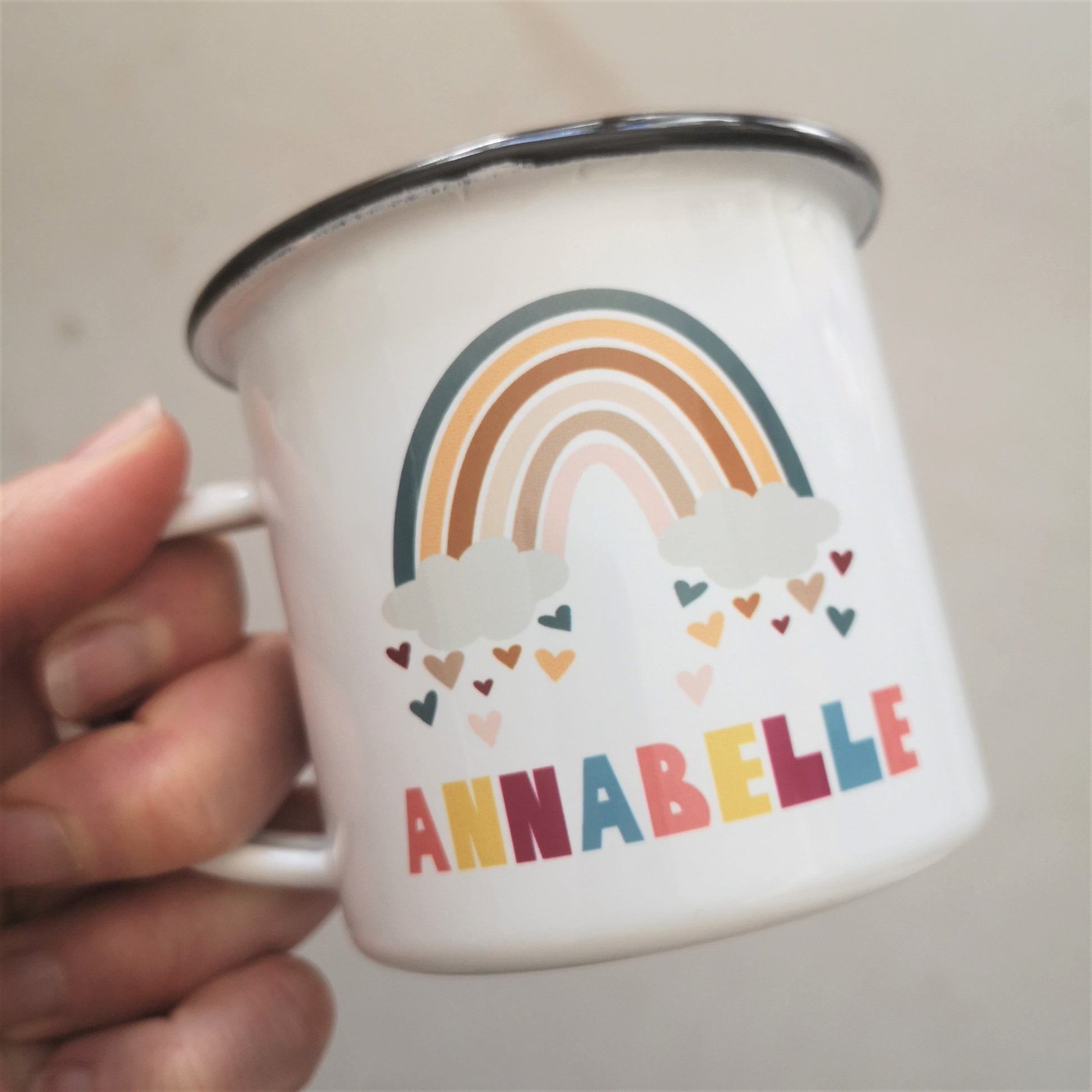 Another angle of a white enamel mug with a cute colourful rainbow on it with hearts just below the clouds, and below the space to add your favourite little one's name.