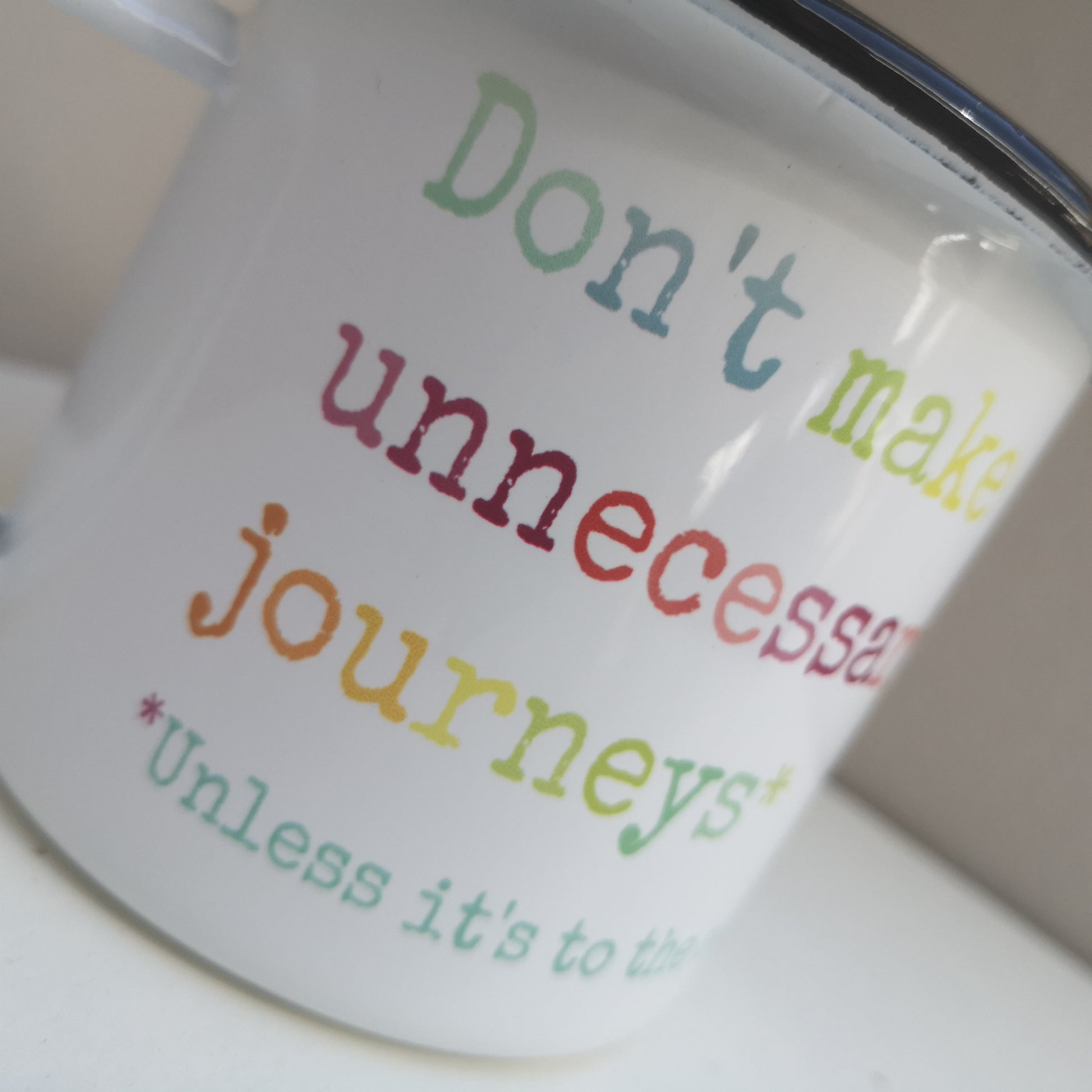 A close up of a white steel enamel mug with the Teresa Mannion catchphrase DON'T MAKE UNNECESSARY JOURNEYS - UNLESS IT'S TO THE SEA written in rainbow coloured type font below