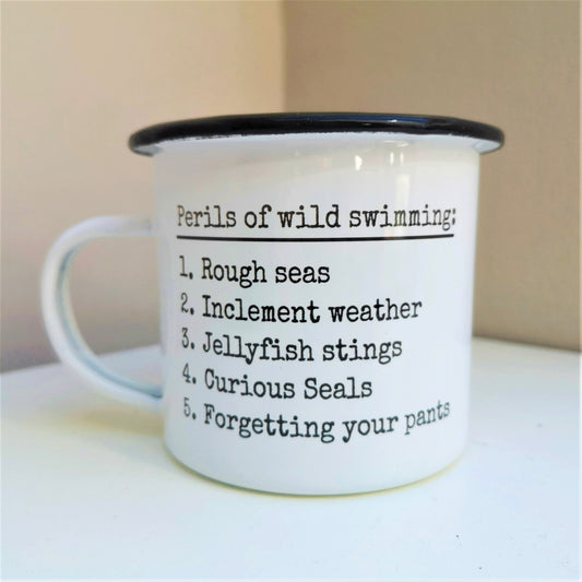 A White enamel mug with a black rim with the following on the front - Perils of wild swimming: Rough seas, Inclement weather, Jellyfish stings, curious seals, Forgetting your pants.