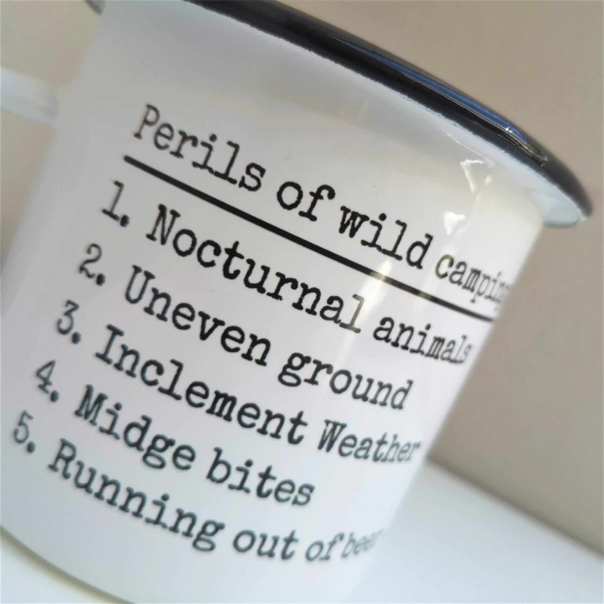 A close up photo of a White enamel mug with a black rim with the following on the front -Perils of wild camping: Nocturnal Animals, Uneven ground, Inclement weather, midget bits, Running out of beer