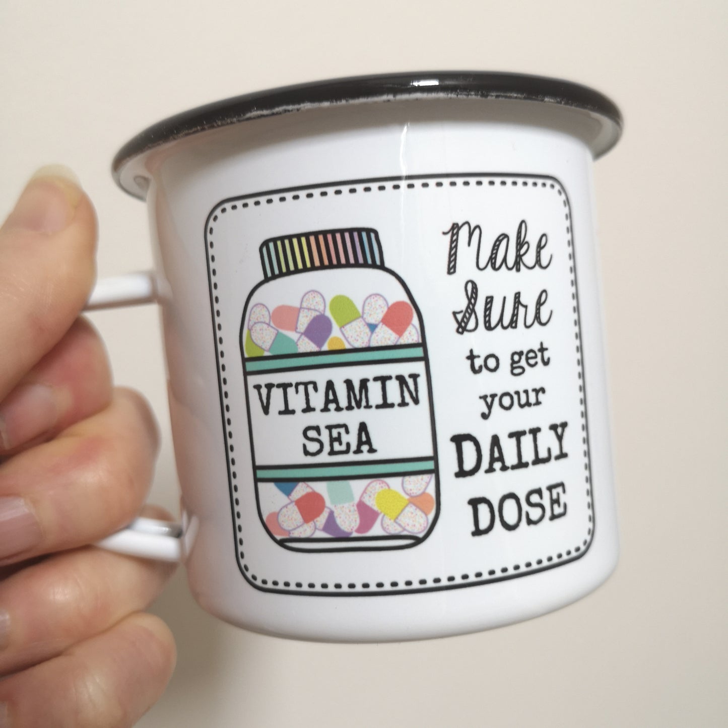 A close up photo of someone holding a White enamel mug with a black rim with the following on the front - a framed hand drawn image of a vitamin jar filled with brightly coloured pills and text to the right of it that says "make Sure to get your daily dose". The bottle has a VITAMIN SEA label on it.