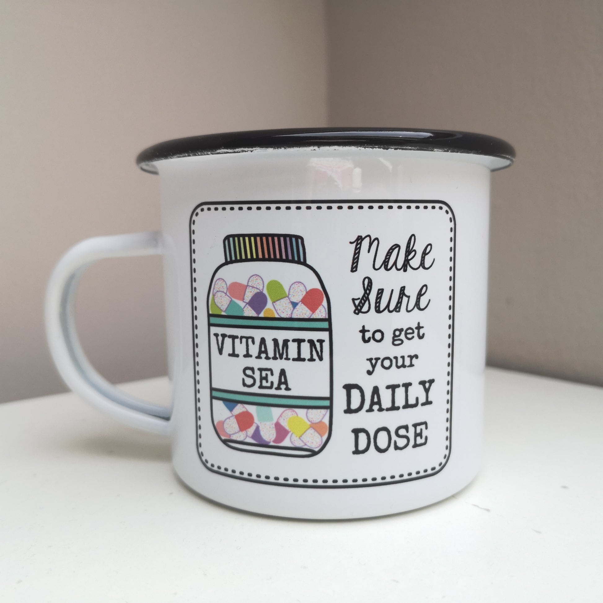 A White enamel mug with a black rim with the following on the front - a framed hand drawn image of a vitamin jar filled with brightly coloured pills and text to the right of it that says "make Sure to get your daily dose". The bottle has a VITAMIN SEA label on it.