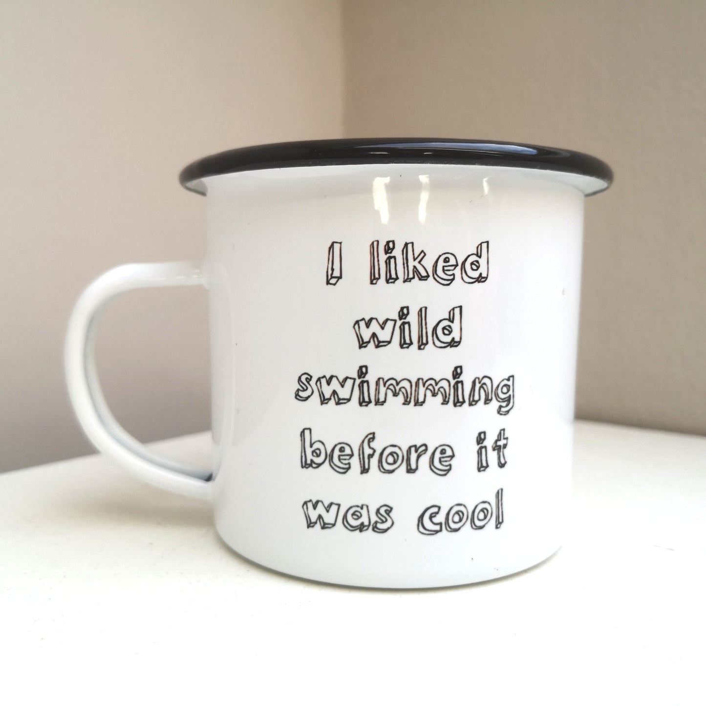 A white steel enamel mug with a black rim, with the following text in black 3d writing - I LIKED WILD SWIMMING BEFORE IT WAS COOL