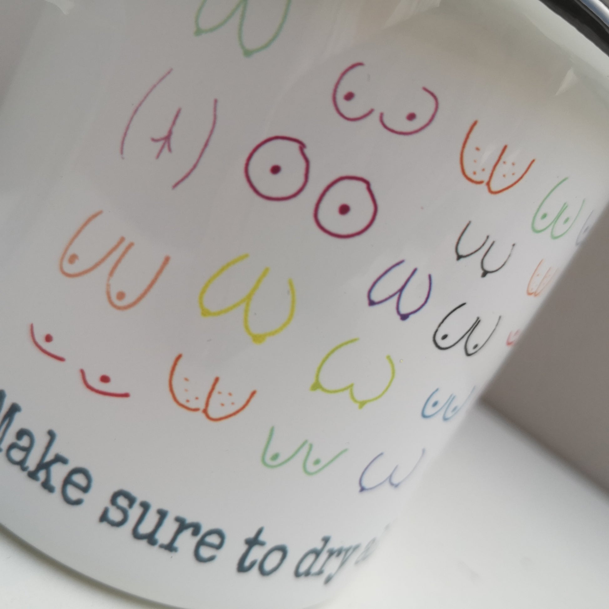 A close up of a  White enamel mug with a black rim with the following design wrapped around the mug  - MAKE SURE TO DRY YOUR WOBBLY BITS AND DON'T FORGET YOUR PANTS and above the text lots of different coloured hand drawn boobs and bums