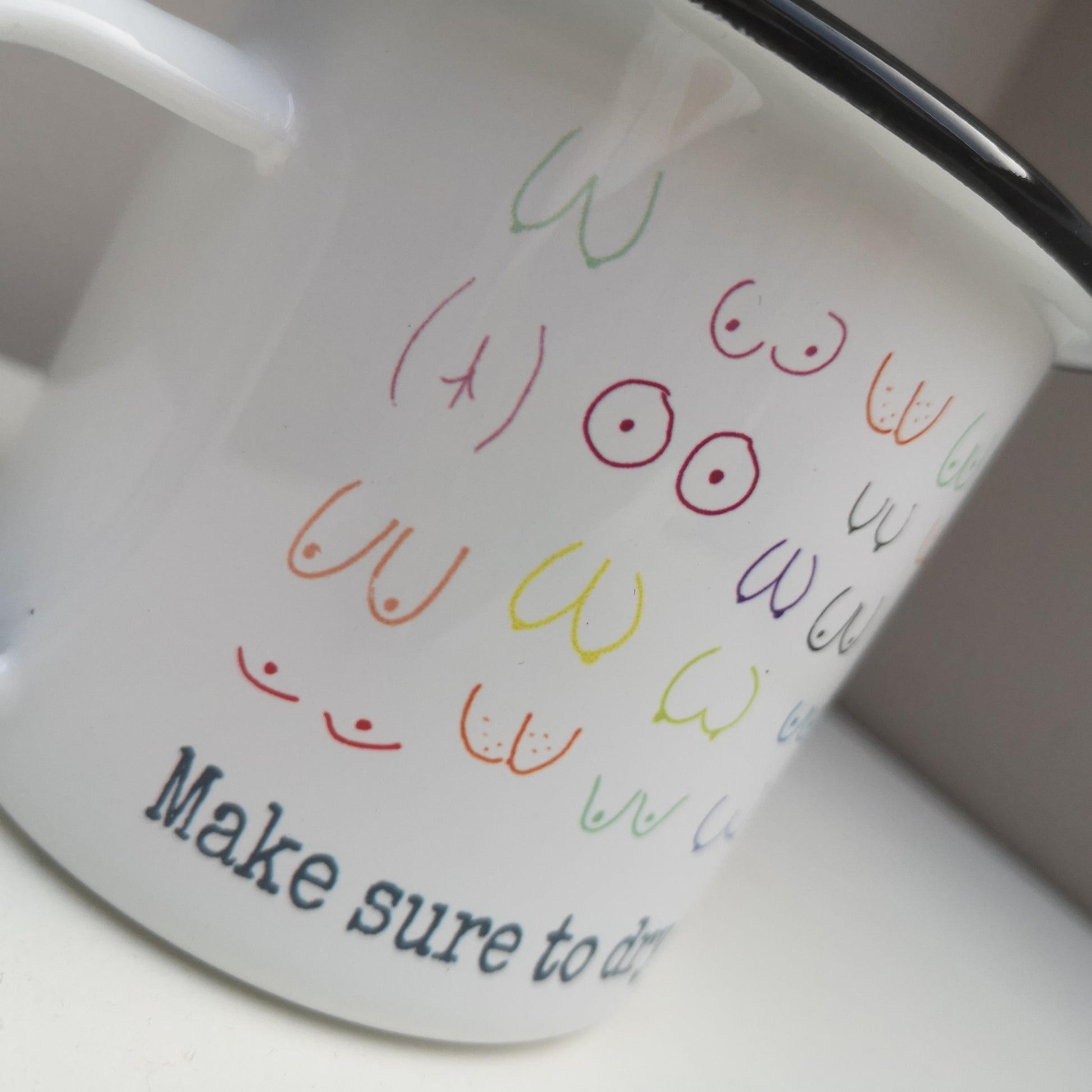 A close up of a White enamel mug with a black rim with the following design wrapped around the mug  - MAKE SURE TO DRY YOUR WOBBLY BITS AND DON'T FORGET YOUR PANTS and above the text lots of different coloured hand drawn boobs and bums
