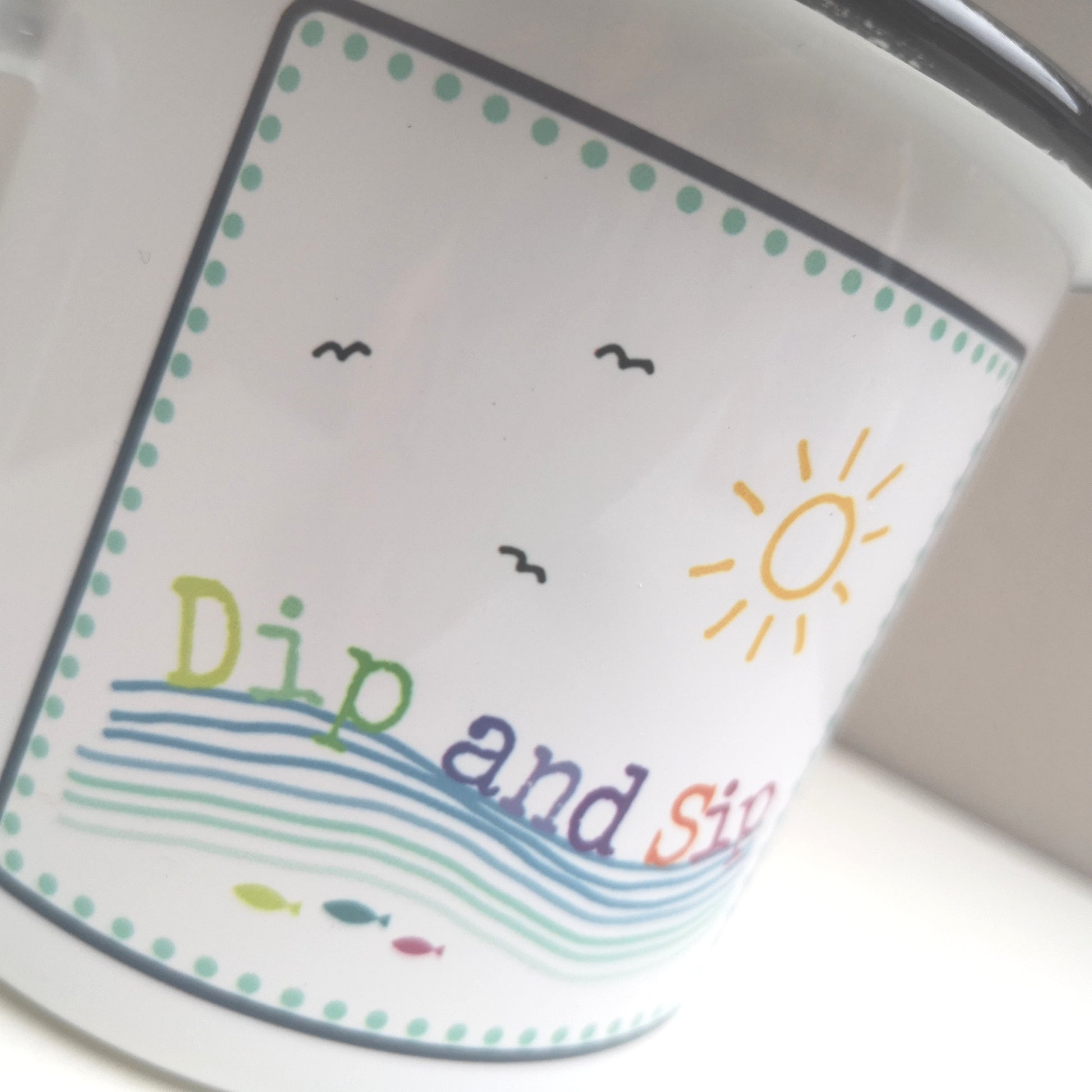 A white steel enamel mug with a year round swimmer's mantra on it - dip and sip. Photo shows a close up of the rainbow coloured design with 3 fish