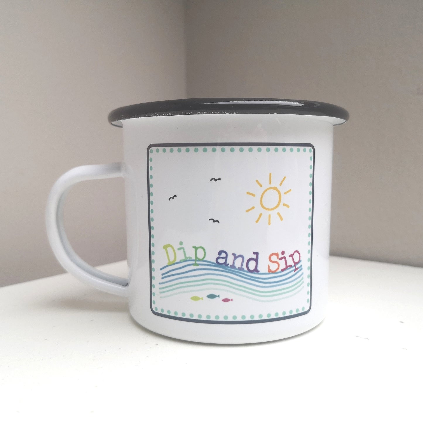 A white steel enamel mug with a year round swimmer's mantra on it - dip and sip. Photo shows the rainbow coloured design with 3 fish