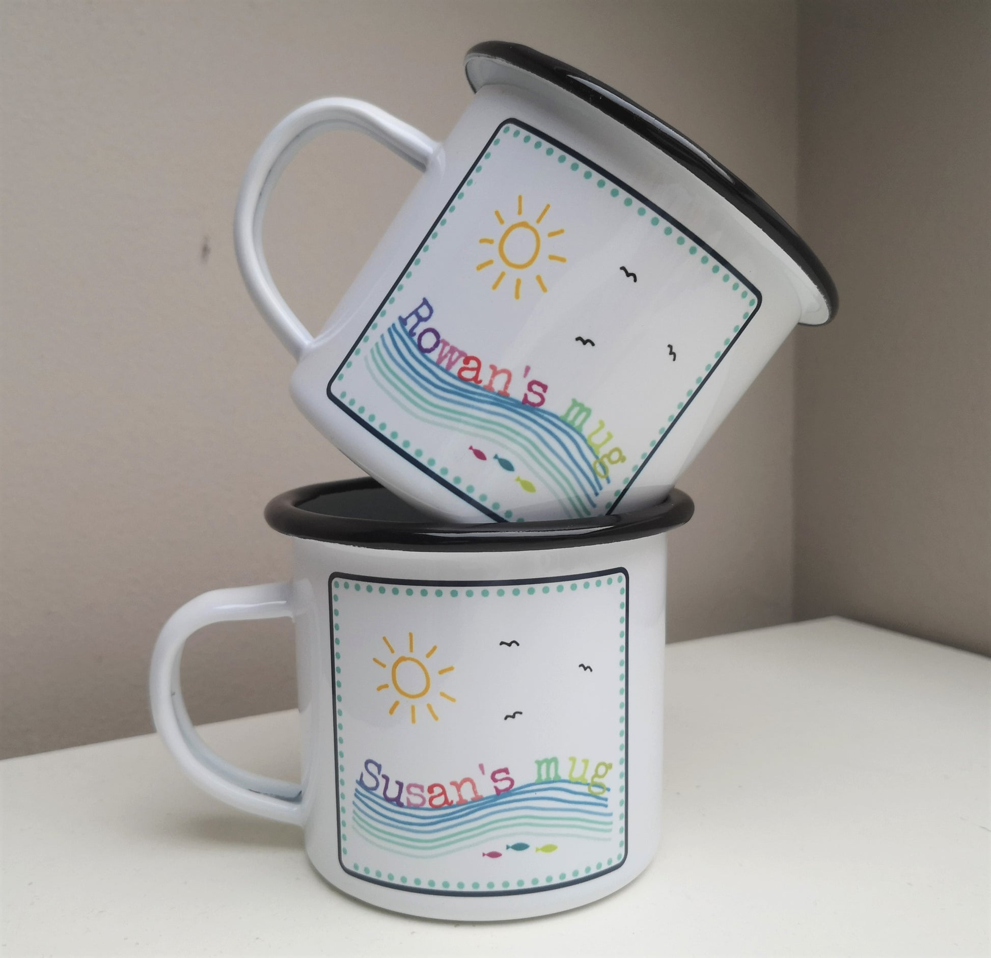 A different view of 2 white enamel mugs that are stacked,  featuring a colourful framed seaside scene, with the mug owner's name riding on the waves!
