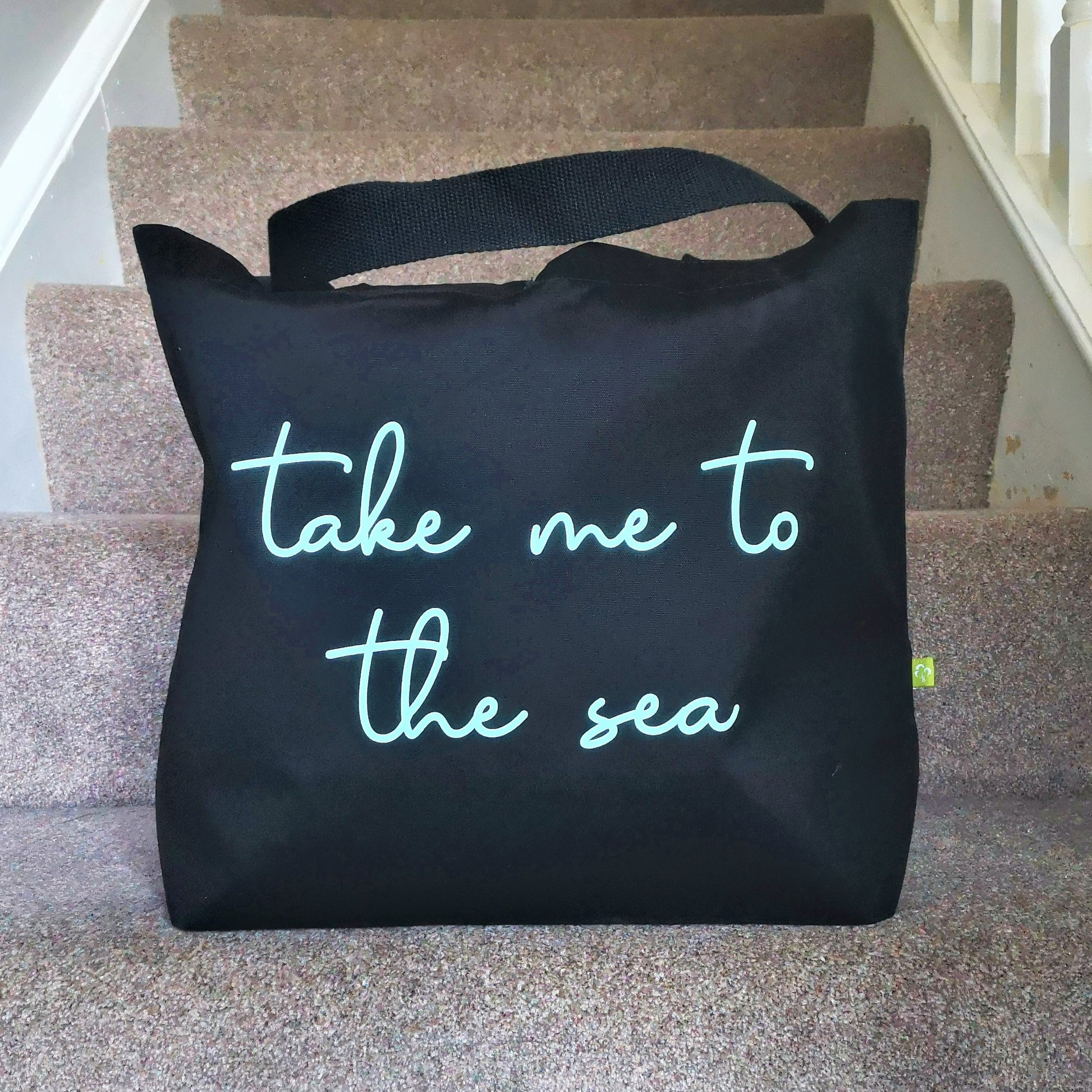 A black organic cotton tote with toggle closing with the following text on the front in mint green type font - Take me to the sea.