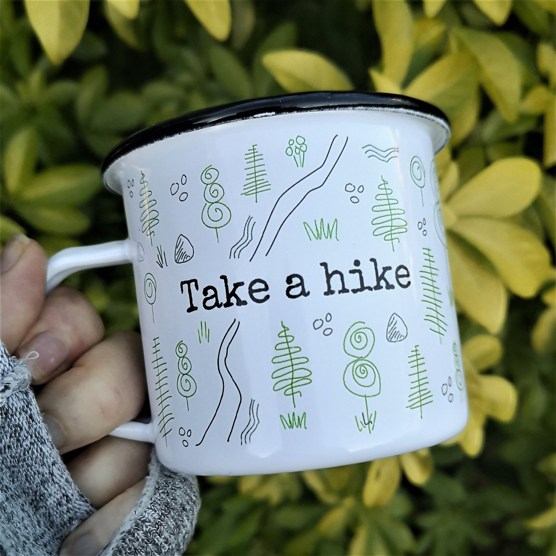 An outside photo of a White enamel mug with a black rim with the following design wrapping around the outside of the mug - a forest scene with a rough path through it and TAKE A HIKE written in the centre on both the back and front of the mug.