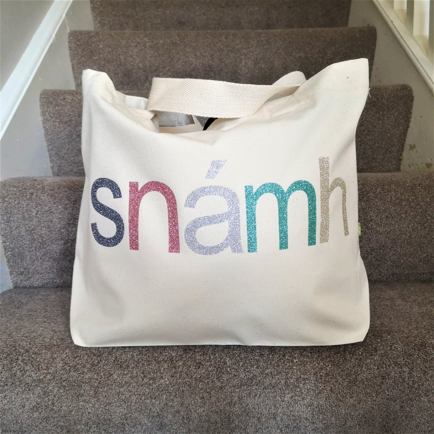  A natural cream organic cotton tote with toggle closing with the following text on the front in various glitter colours in type font - SNÁMH