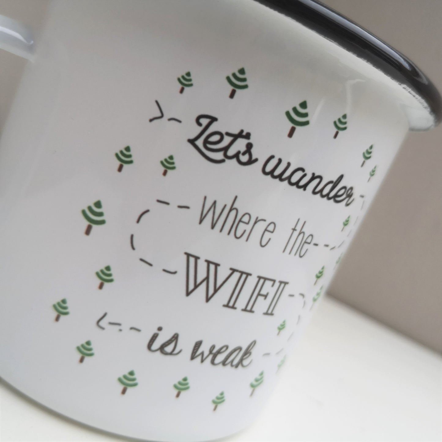 A close up of a White enamel mug with a black rim with the following on the front - 'Let's wander where the WIFI is weak' and lots of little trees that are upside down wifi signals