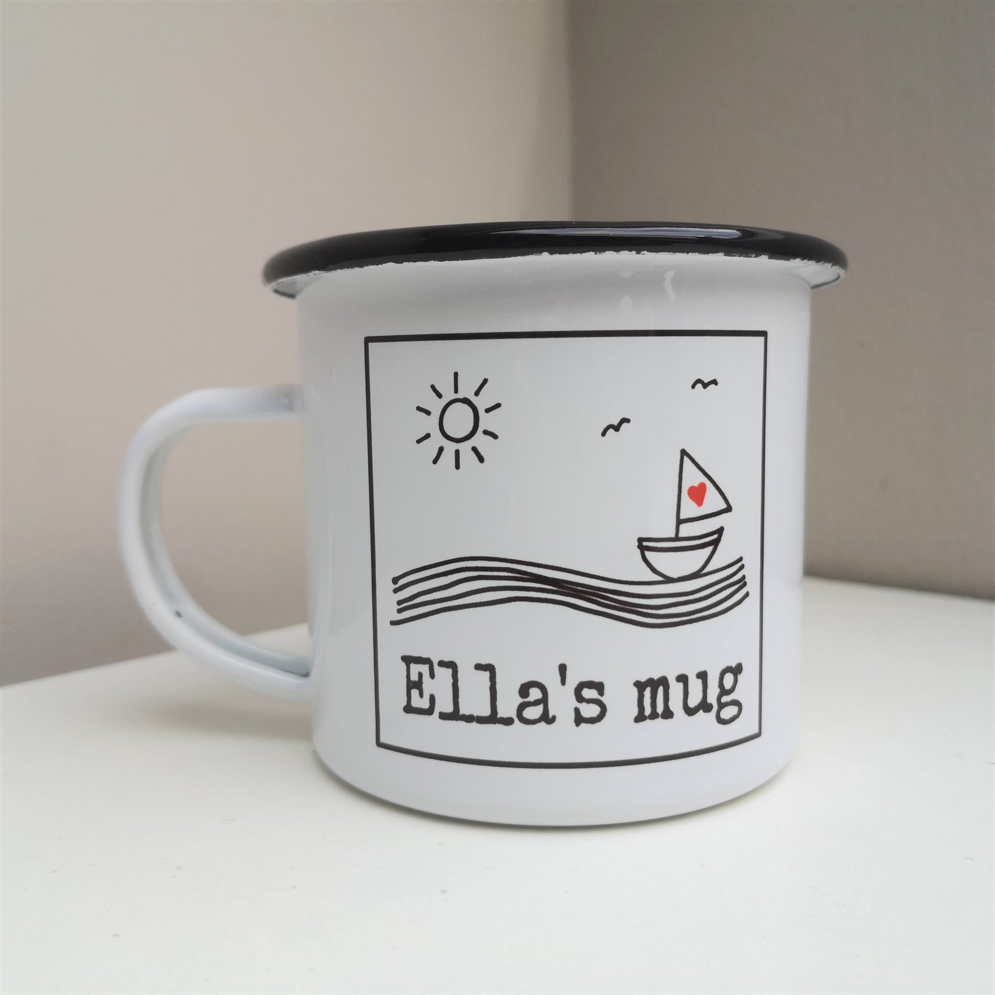 A white enamel mug with a black & white framed hand drawn seaside scene, with space along the bottom for the owner's name.