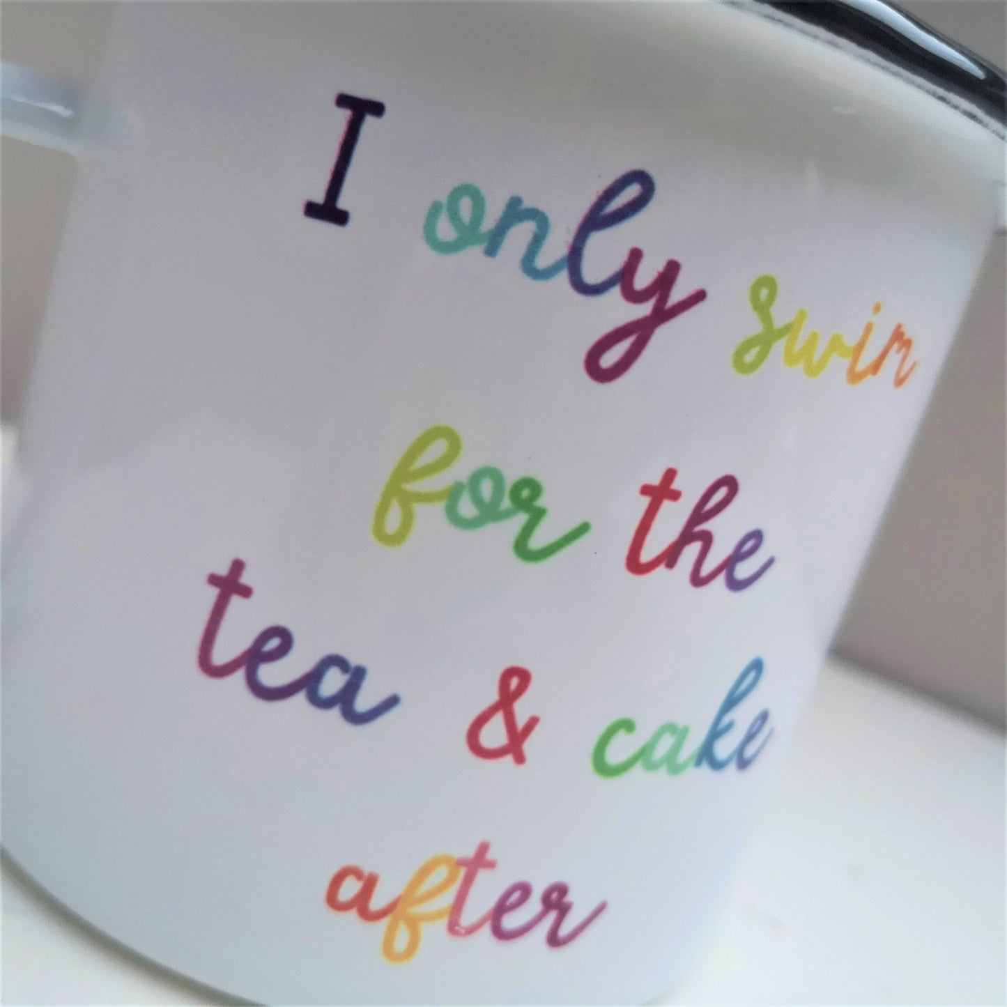 A close up of a steel enamel cup for your after-swim tipples, with a choice of 3 confessions in colourful rainbow text.  This mug has the following excuse - I only swim for the tea & cake after.  