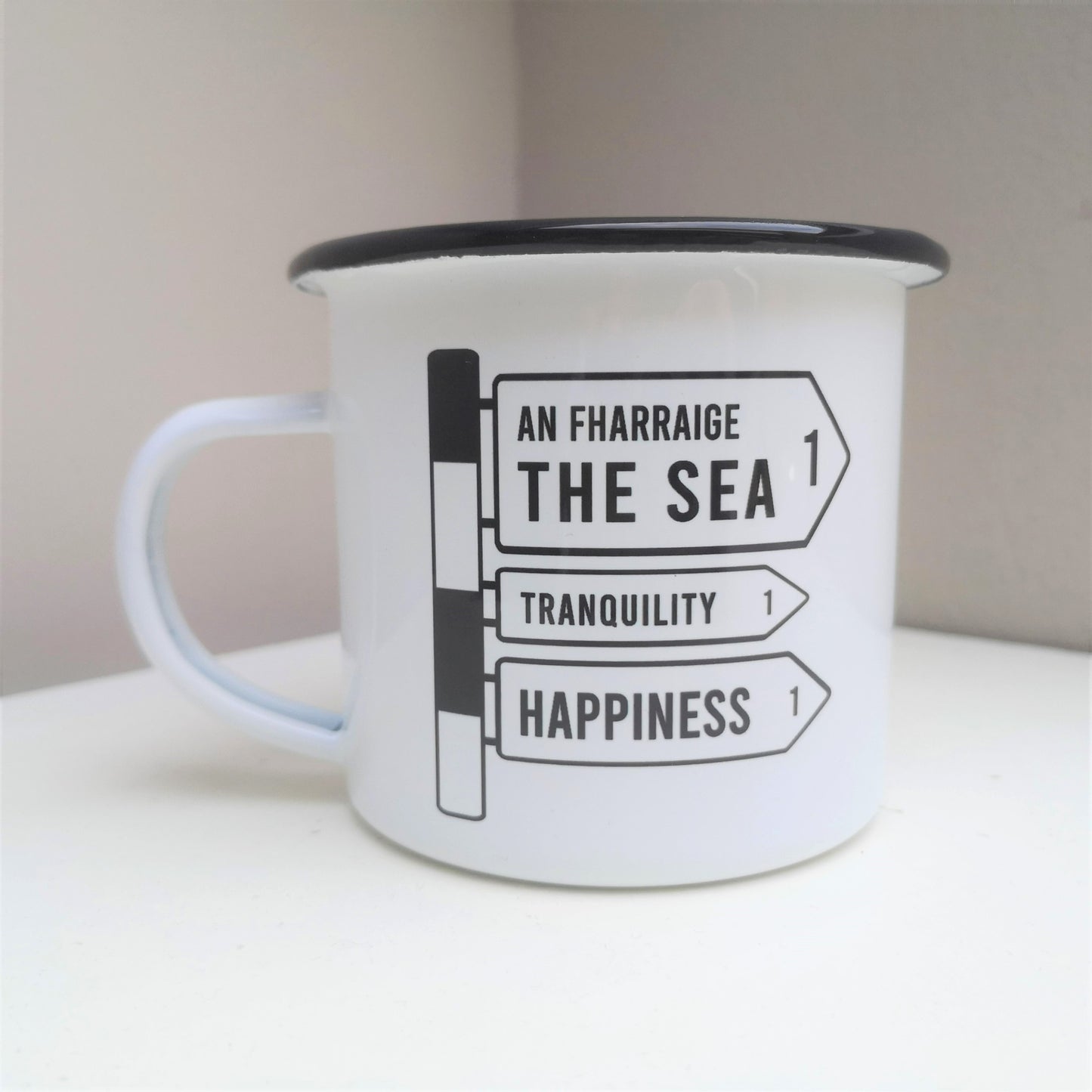 A White enamel mug with a black rim with the following on the front -an old Irish Roadsign pointing to the right with arrows for The Sea, Tranquility and Happiness