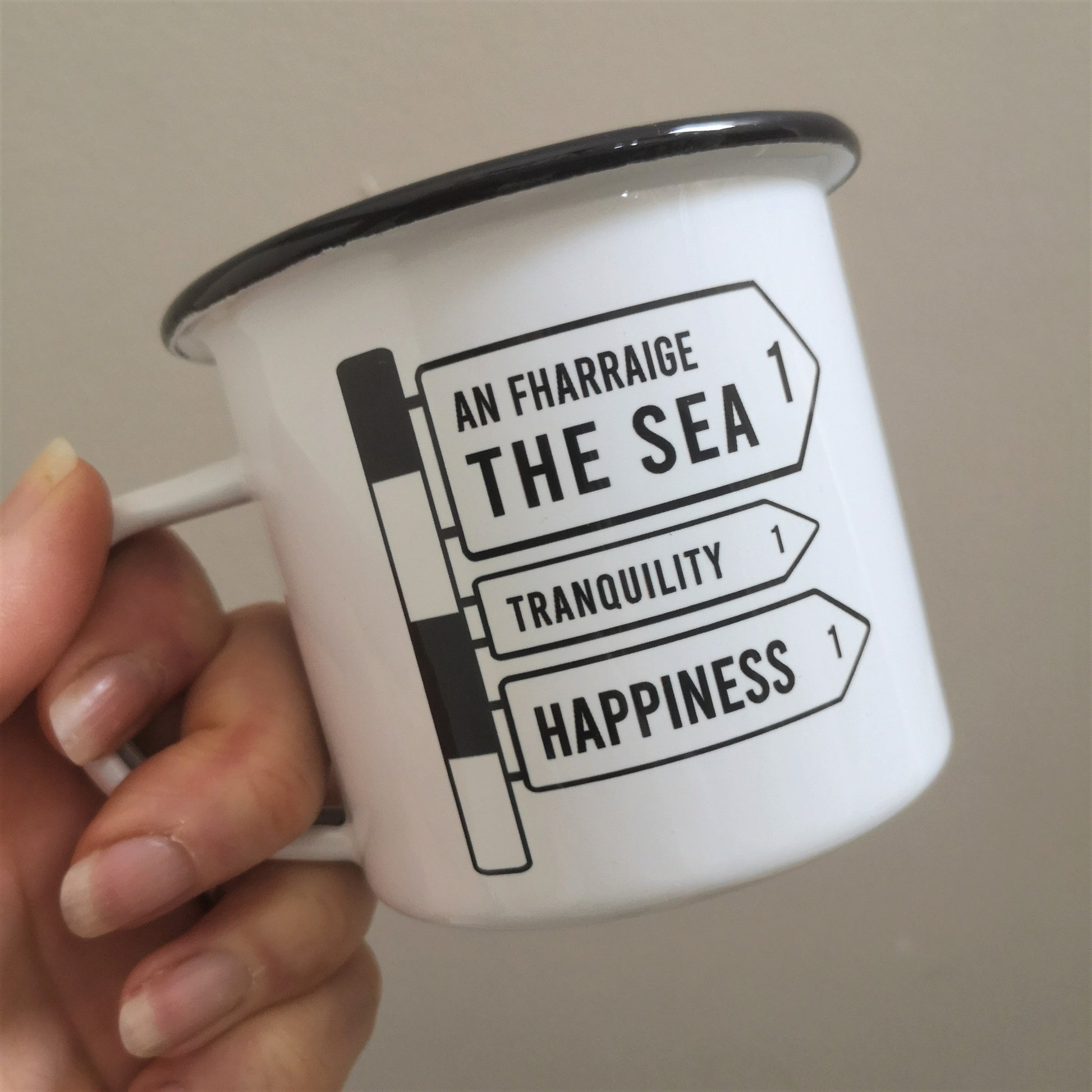 A different angle of a White enamel mug with a black rim with the following on the front -an old Irish Roadsign pointing to the right with arrows for The Sea, Tranquility and Happiness