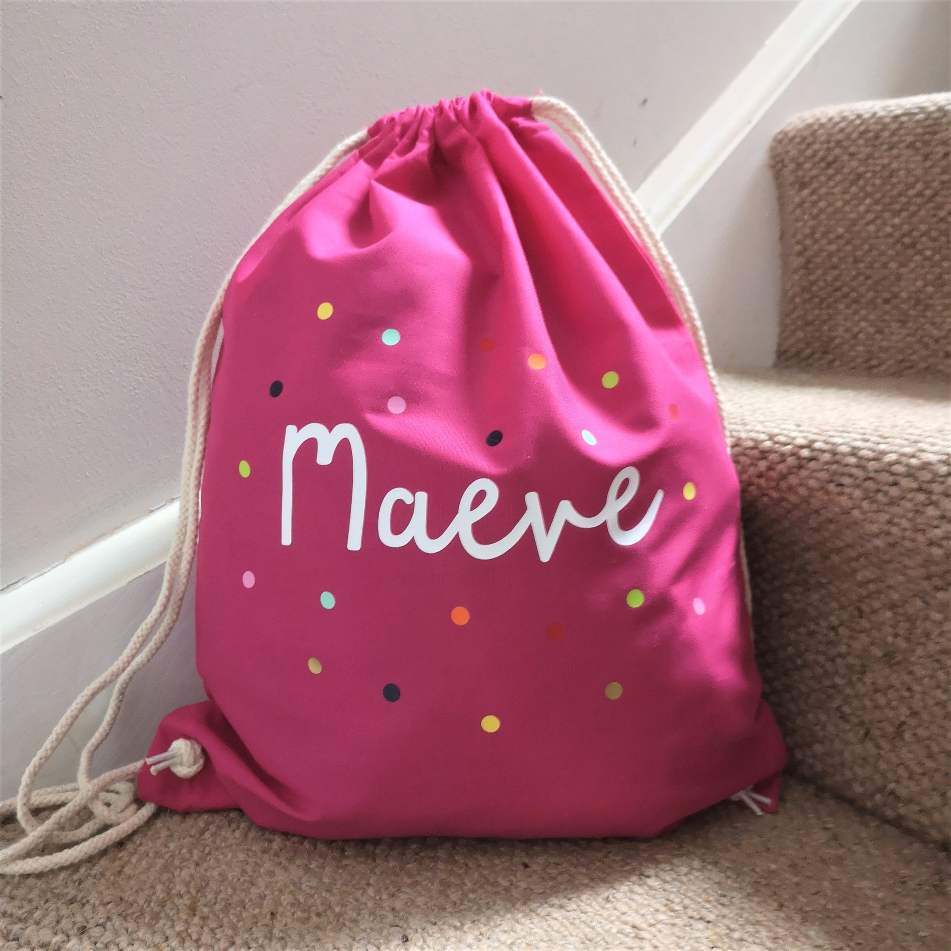 A photo showing the pink bag colour choice of a personalised light cotton drawstring bag with dots and personalised with a name.  The 3 colour choices are french navy, fuschia pink and light grey