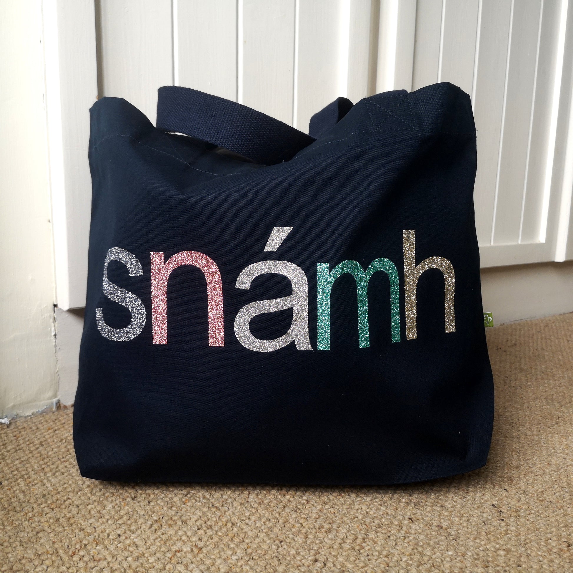  A navy organic cotton tote with toggle closing with the following text on the front in various glitter colours in type font - SNÁMH