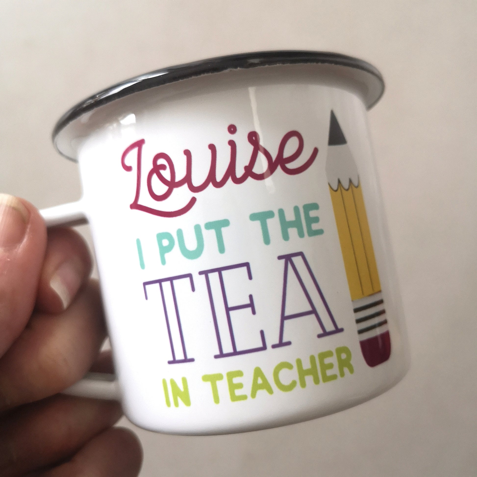 A close up of a White enamel mug with a black rim with the following on the front - <NAME> I out the tea in teacher and a big pencil to the right of the text