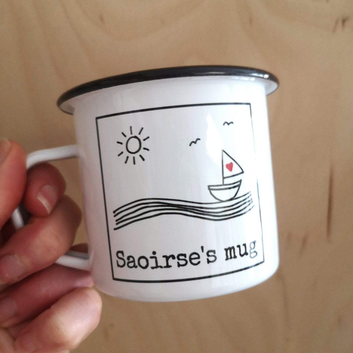 A different view of a white enamel mug with a black & white framed hand drawn seaside scene, with space along the bottom for the owner's name.