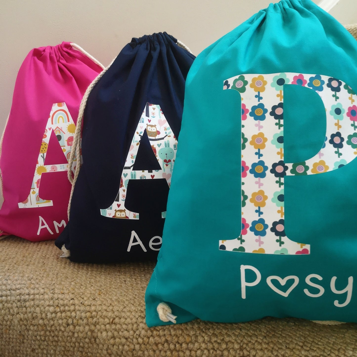 A photo showing 3 of the bag colour choices for A personalised light drawstring bag with a patterned Initial and their name underneath