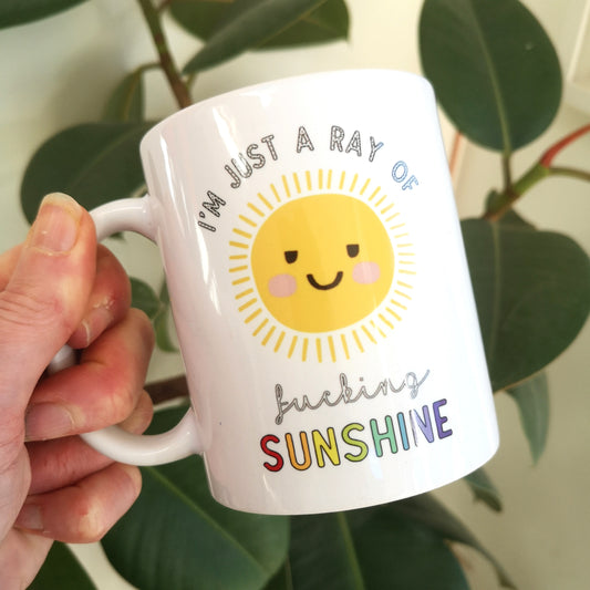 A white ceramic mug with a bright picture of a happy sun on it, surrounded with the following text in rainbow coloured writing - I'm just a ray of f*cking Sunshine