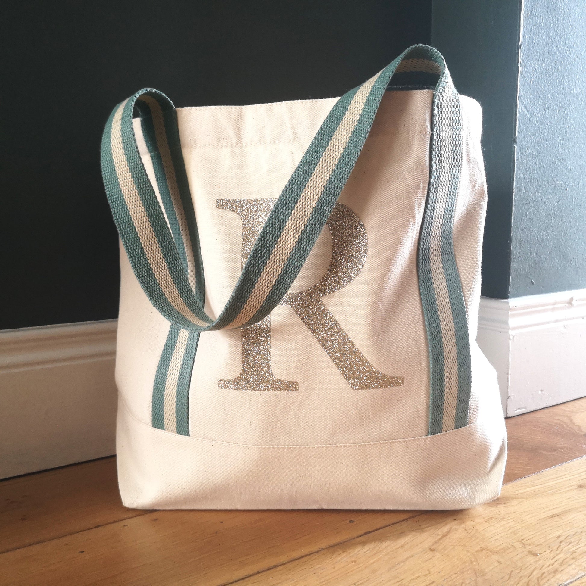 A sturdy natural organic cotton tote with sage green and gold striped straps, and a glittery gold initial on the front in large type