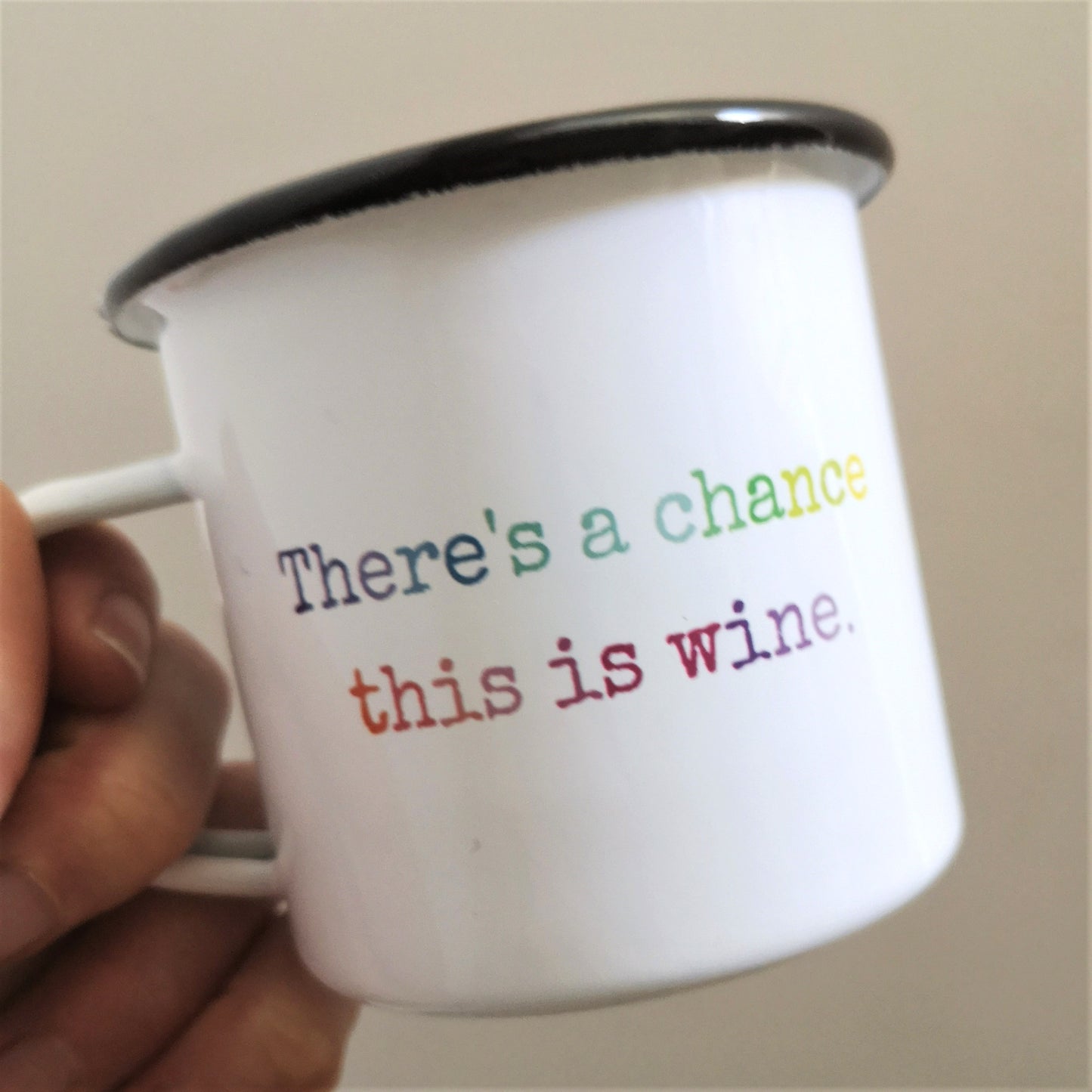 A close up of a hand holding a White enamel mug with a black rim with the following on the front in rainbow lettering - there's a chance this is wine