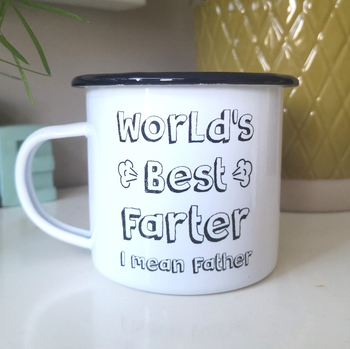 A White enamel mug with a black rim with the following on the front - World's Best Farter, I mean Father
