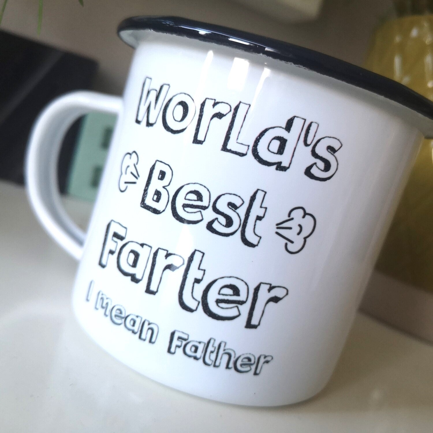 A close up of a White enamel mug with a black rim with the following on the front - World's Best Farter, I mean Father