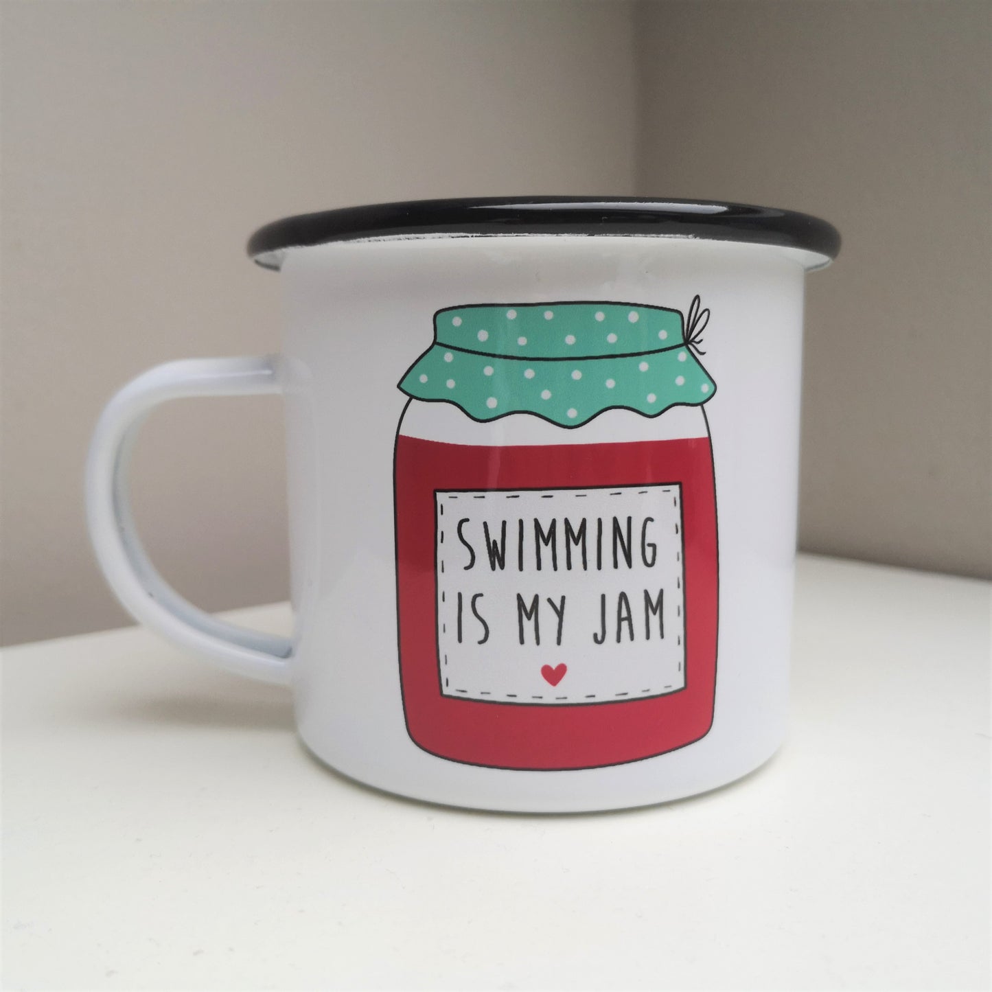 A White enamel mug with a black rim with a colourful red and green jam jar on the front, containing the following text - SWIMMING IS MY JAM
