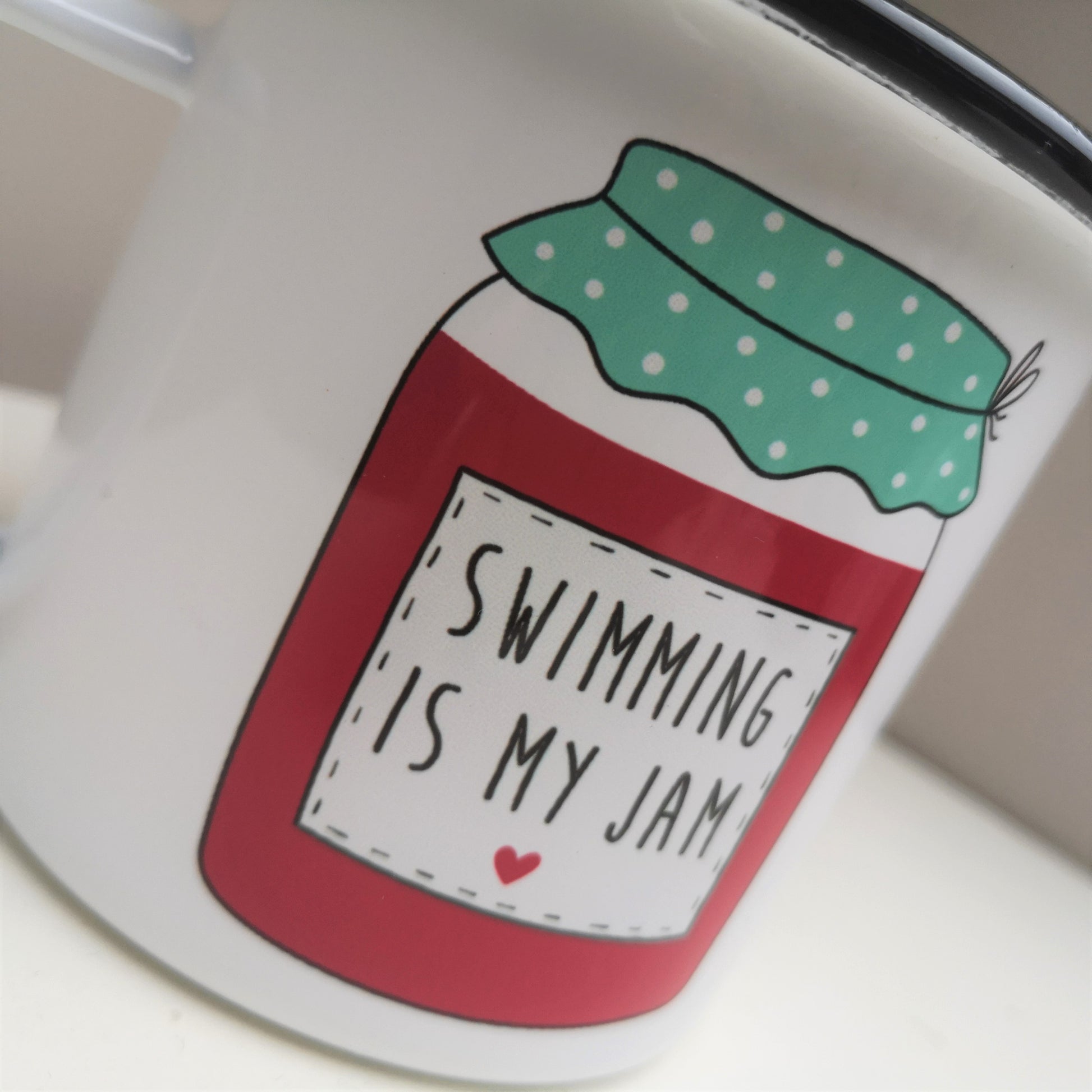 A close up view of a White enamel mug with a black rim with a colourful red and green jam jar on the front, containing the following text - SWIMMING IS MY JAM