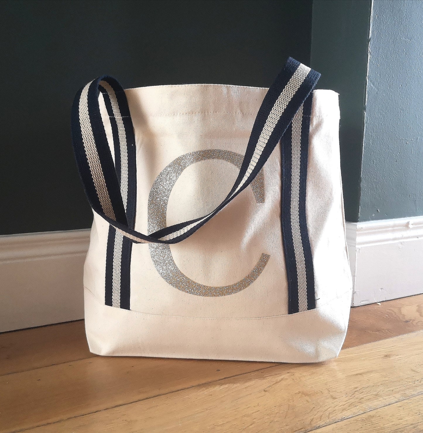 A sturdy natural organic cotton tote with navy and gold striped straps, and a glittery gold initial on the front in large type
