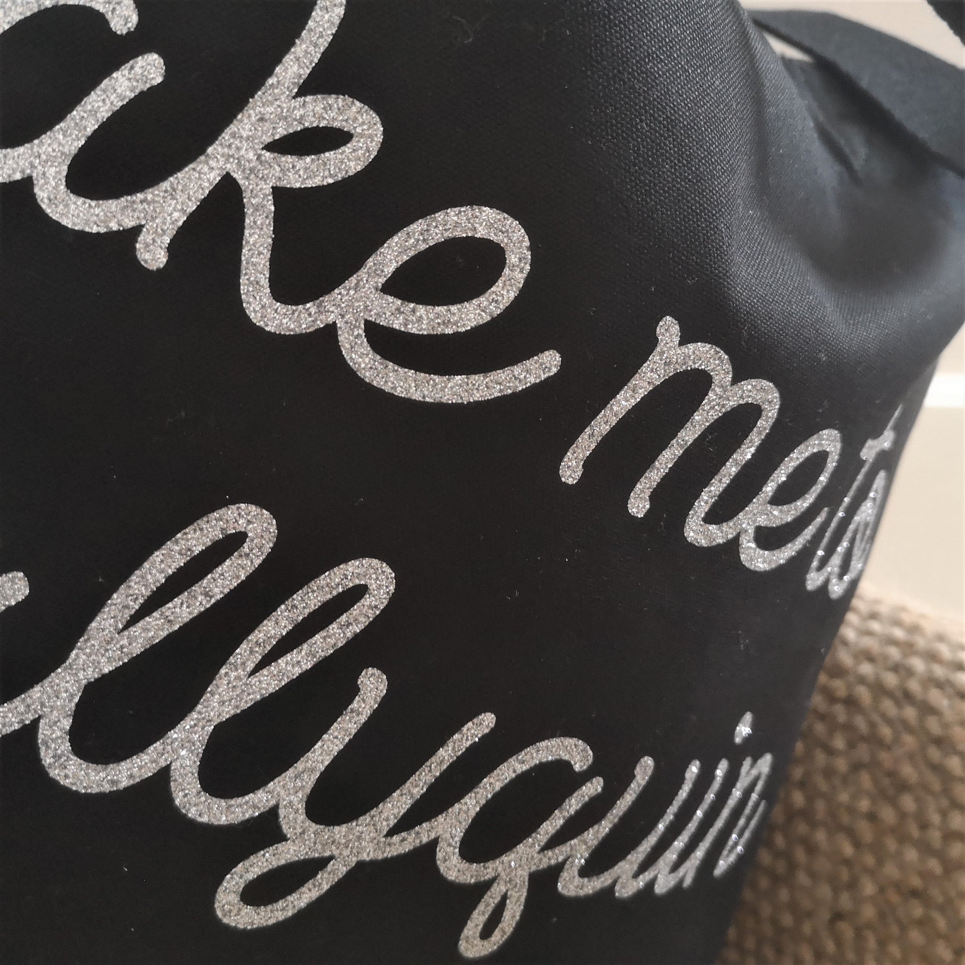A close up of a black organic cotton tote  with toggle closing with the following text on the front in silver glittery swirly font - Take me to (your choice of text here)