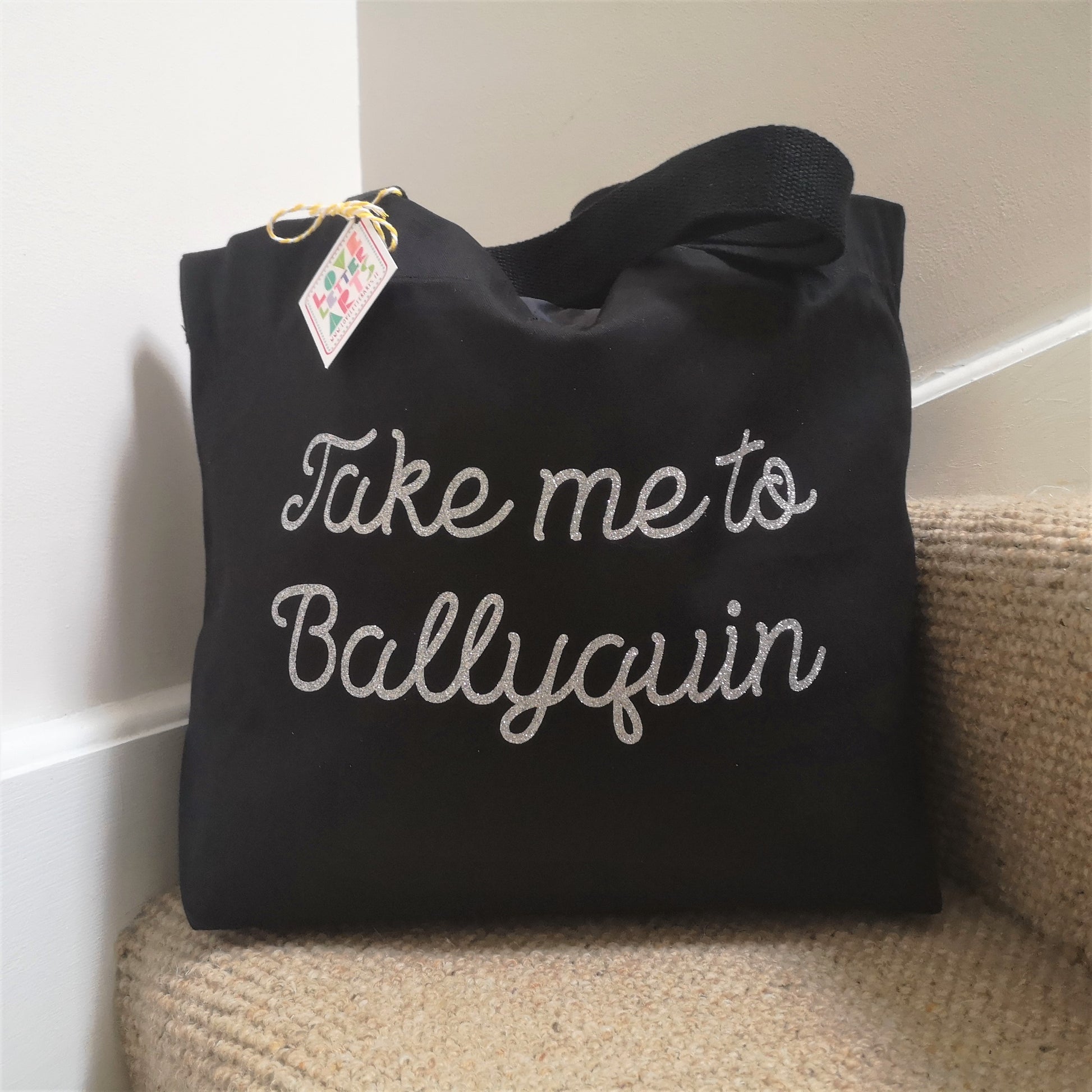 A black organic cotton tote with toggle closing with the following text on the front in white type font - Take me to (your choice of text here). This one is in silver glitter text.